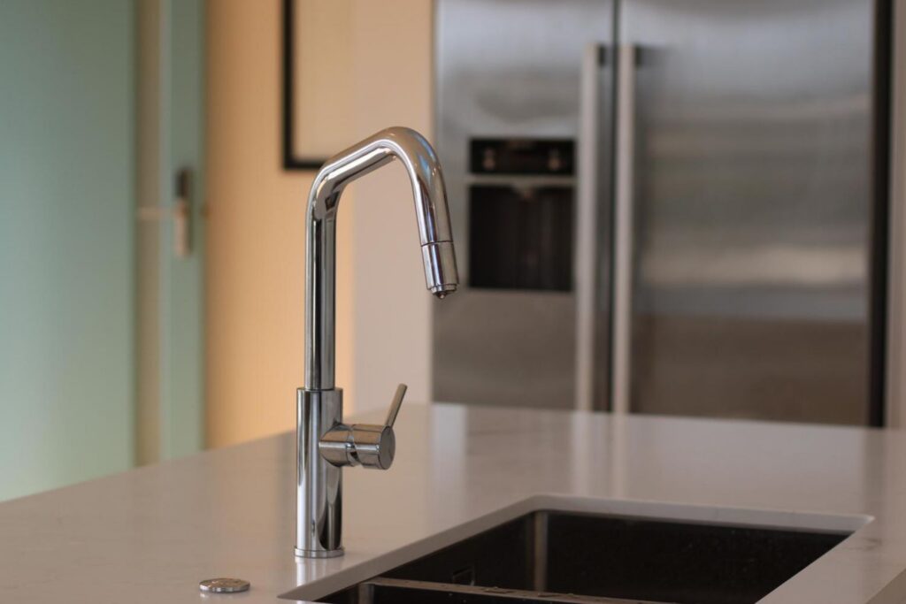 A modern chrome kitchen faucet extends over a white countertop with a stainless steel refrigerator in the blurred background.