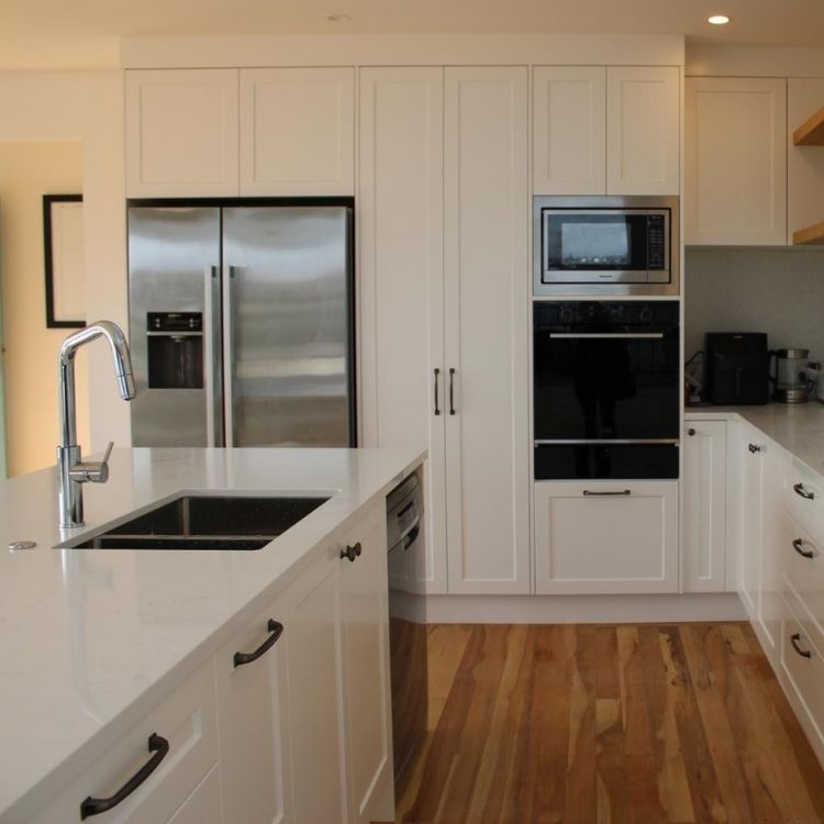 A modern white kitchen with a stainless steel refrigerator, built-in oven and microwave, a large kitchen island with a sink, and wooden floors.