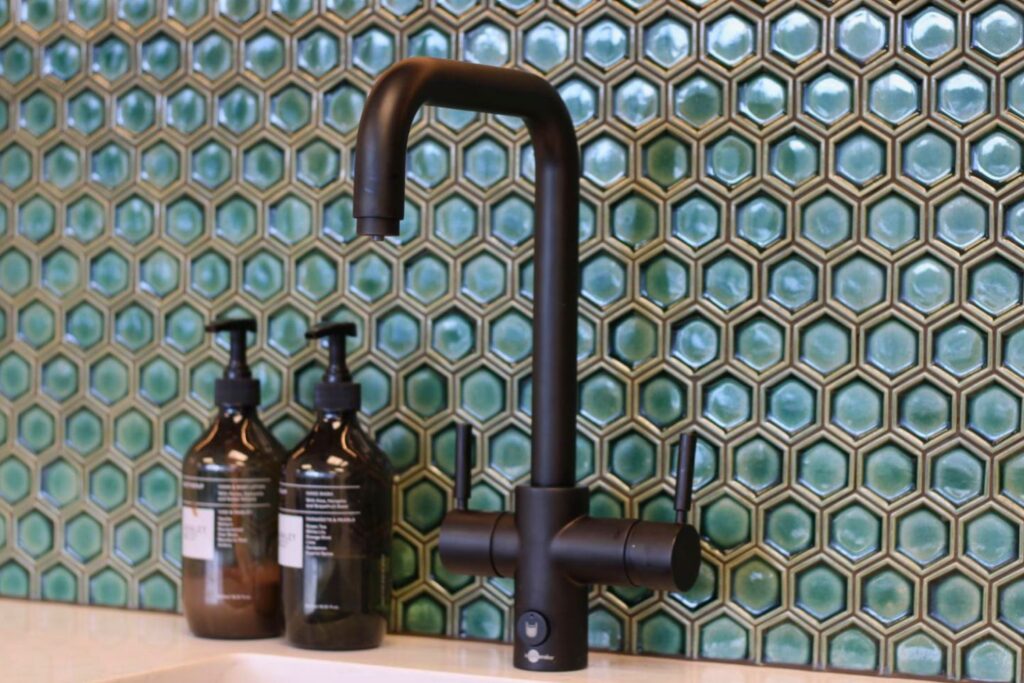A modern black faucet stands in front of a wall tiled with hexagonal green and gold tiles. Two dark amber soap dispensers are to the left of the faucet. Great kitchen renovation.