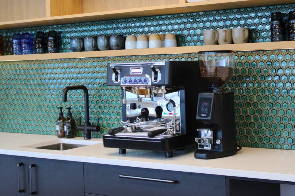 A professional plumbed-in espresso machine and coffee grinder sit on a counter in front of a teal hexagonal tile backsplash. There is a shelf above with different cups and a sink with faucet to the left of the coffee machine. Great kitchen renovation.