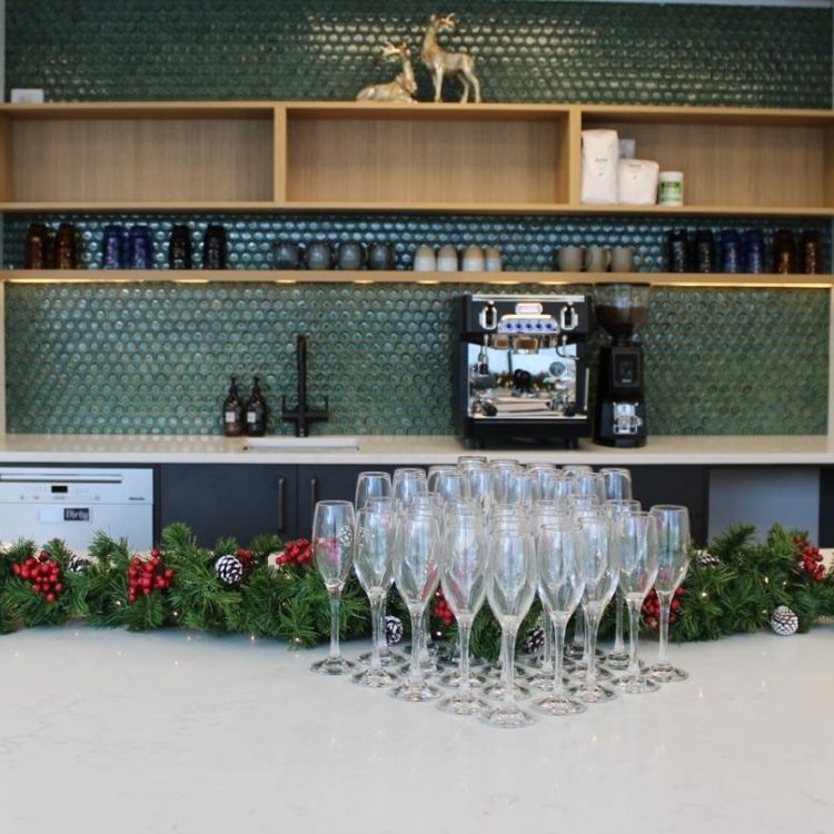A bar area with a coffee machine, shelves stocked with mugs and bottles, and champagne flutes arranged on the counter, decorated with a festive Christmas garland. Great office kitchen renovation.