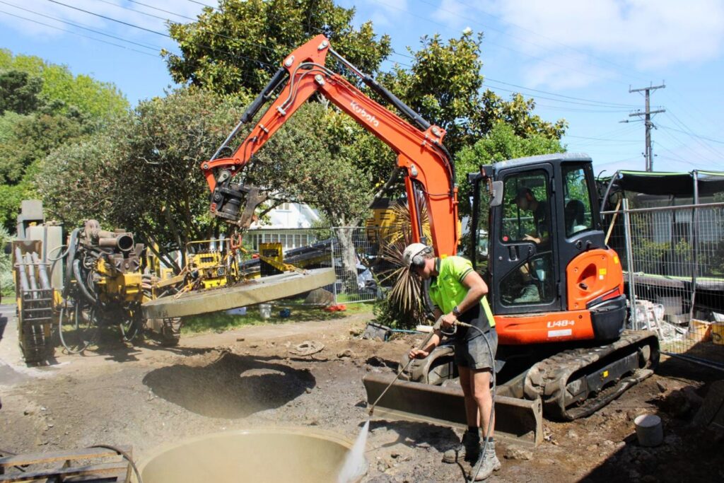 An orange excavator is digging lifting up a plate from a drainage chamber while a worker in a neon vest sprays water with a hose.