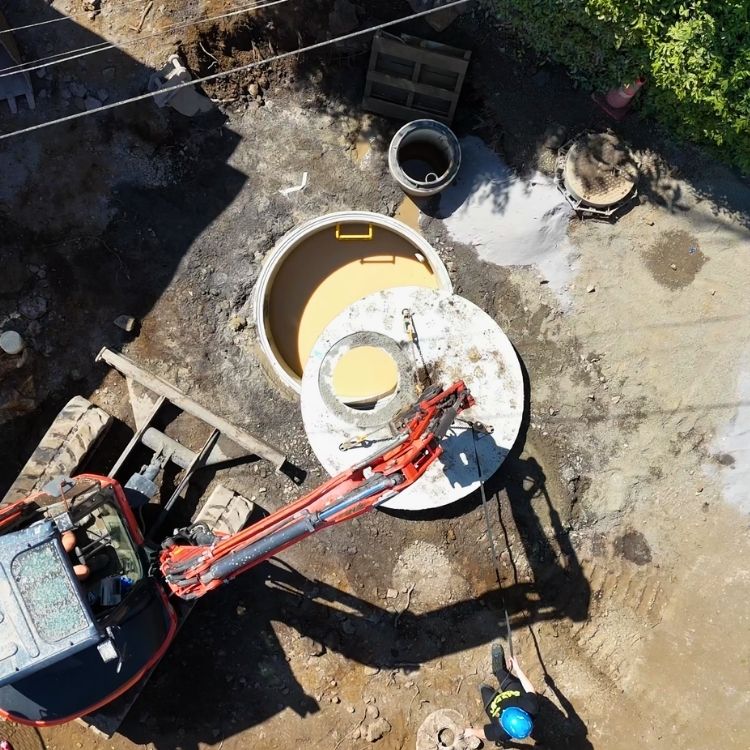 An aerial view of construction site with an excavator lifting a concrete plate onto a drainage chamber. A worker in a blue helmet stands nearby guiding the plate.