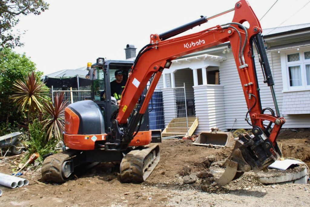 An orange Kubota excavator is digging at a construction site next to a white house, with a person operating it. There are white pipes in the left corner and a drainage chamber in the background.